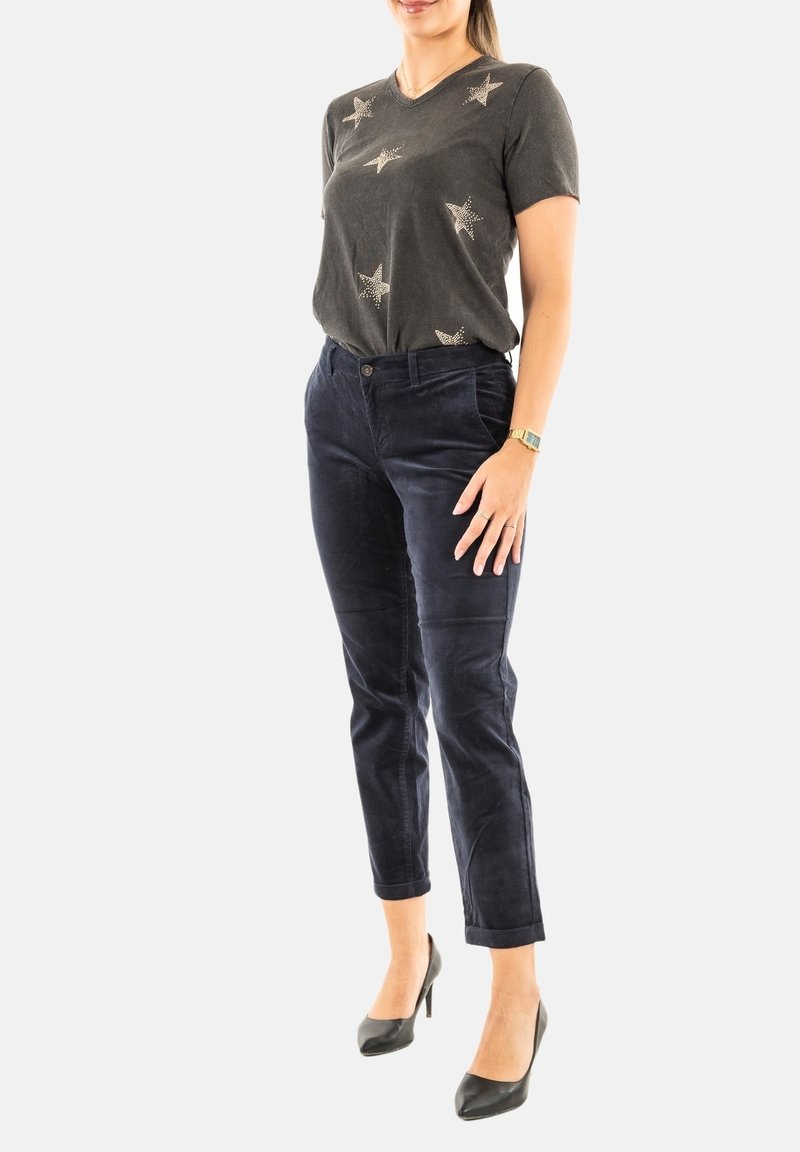 Femme portant une chemise à manches courtes sombre à motifs d'étoiles, un pantalon slim bleu marine, des talons hauts noirs et une montre en or, posant devant un fond blanc.