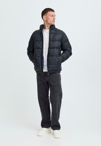Svart pufferjacka med quiltat mönster, hög krage och dragkedja. Buren över en vit skjorta, kombinerad med mörka jeans och vita sneakers.