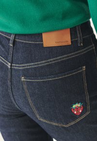 Tummansiniset farkut sisältävät nahkapaikan, jossa lukee "TANTUUM" vyötäröllä, sekä värikkään brodeeratun mansikkakuosin takataskussa.