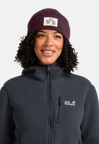 Femme souriante avec des cheveux bouclés portant un bonnet en tricot Jack Wolfskin violet foncé et une veste polaire noire zippée avec un logo sur la poitrine.