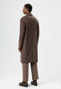 Long manteau en laine marron avec col cranté, fente unique à l'arrière, et poignets boutonnés, associé à un pantalon beige et des chaussures foncées.