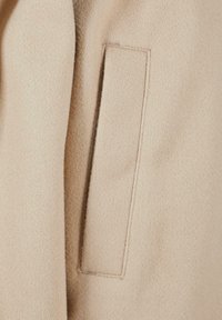 Beige ullfrakk med en jevn tekstur, rektangulær lomme og minimale sømdetaljer. Myk, varm og skreddersydd design.