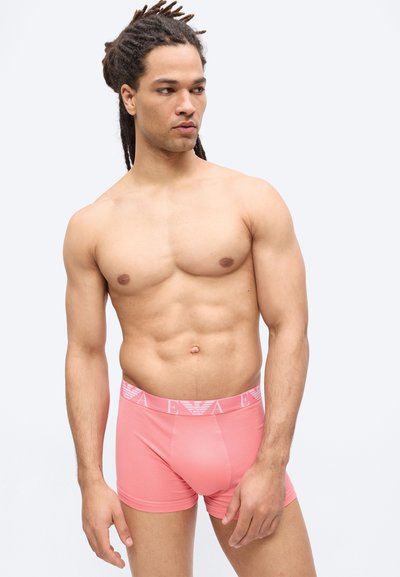 Ένας γυμνασμένος άντρας με κολιές, φορώντας ροζ μπόξερ με λογότυπο στη μέση, στέκεται μπροστά σε απλό φωτεινό φόντο, κοιτώντας πλάγια.