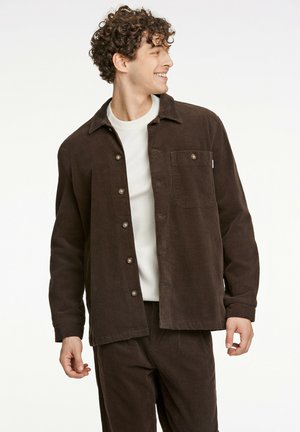 Brown corduroy shirt jacket with a collar, front buttons, and one chest pocket, worn over a light sweater and paired with matching pants.