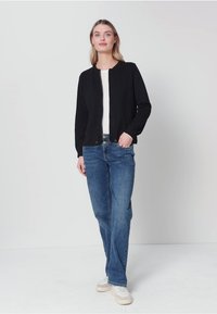 Kobieta stojąca z jedną ręką w kieszeni, ubrana w czarną kurtkę na zamek, białą koszulę, niebieskie dżinsy i białe trampki na jednolitym tle.