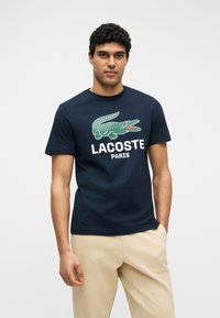 Άνδρας που φοράει μπλουζάκι μπλε ναυτικό με πράσινο λογότυπο κροκόδειλου και κείμενο "LACOSTE PARIS", σε συνδυασμό με μπεζ παντελόνι, στέκεται μπροστά σε λευκό φόντο.