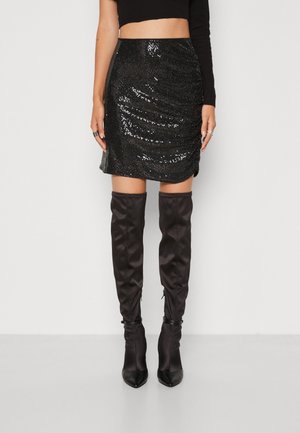 Vrouw in een zwarte, glinsterende minirok, gecombineerd met zwarte hoge laarzen en een cropped zwart langemouwen shirt, staande tegen een witte achtergrond.