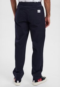 Mørkeblå bukser med elastisk talje og to baglommer, lavet af letvægtsstof. Båret med sorte sneakers med røde detaljer.