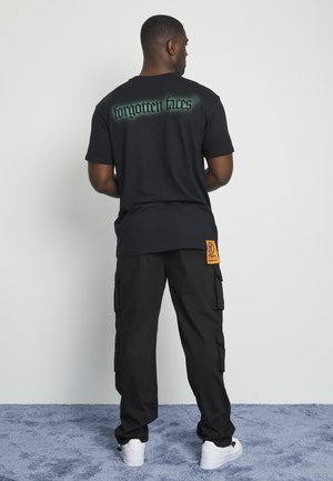 Black cotton t-shirt featuring "forgotten faces" graphic in green on the back, paired with loose black cargo pants and white sneakers.