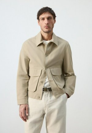 Uomo con giacca beige con bottoni e grandi tasche frontali, camicia bianca, pantaloni bianchi e cintura nera su sfondo chiaro uniforme.