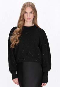 Pulover negru, scurt, realizat dintr-un material moale, cu stropi strălucitori; are un decolteu rotund și mâneci lungi, lejere.