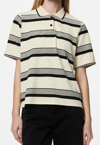 Modell klädd i en kortärmad, randig polo t-shirt i kräm och svart, ihop med svarta byxor, står mot en enfärgad bakgrund.