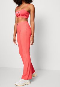 Kobieta w koralowo-różowych, prążkowanych, rozszerzanych spodniach oraz pasującym, krótkim topie z logo, w białych i beżowych sneakersach, stojąca bokiem.