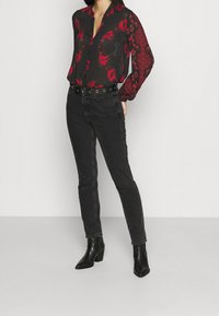 Kobieta w czarno-czerwonej bluzce w wzory, czarnych jeansach o wysokim stanie z paskiem oraz czarnych botkach na obcasie, stoi na białym tle.