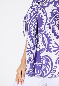 Top morbido in tessuto bianco con un audace motivo paisley viola. Presenta legami laterali e un orlo arrotondato per una vestibilità rilassata.