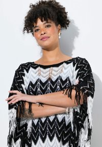 Svart och vit poncho med zigzag-mönster och fransar i kanterna. Tillverkad av lätt stickad tyg, med öppen halsringning och sidosömmar.