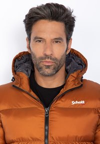 Giacca isolante marrone, morbida e gonfia, con rivestimento interno grigio, chiusura con zip e cappuccio regolabile. Presenta un piccolo logo sul petto.