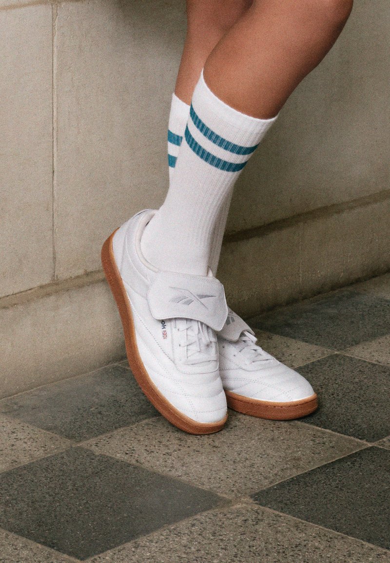 Λευκά sneakers από δέρμα με καουτσούκ σόλα, που διαθέτουν ιμάντα Velcro και λεπτομέρεια λογότυπου. Φορεμένα με λευκές κάλτσες που εμφανίζουν τυπώματα σε τυρκουάζ λωρίδες.