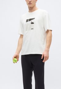 Fehér pamut póló, grafikai mintával szürke és fekete árnyalatokkal, valamint a "BOSS" logóval, sötét nadrággal és zöld labdával párosítva.