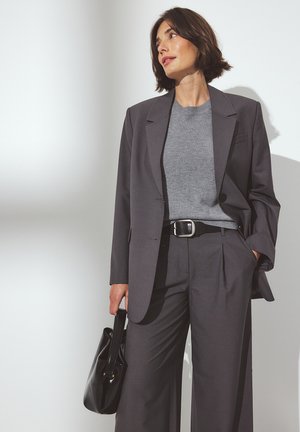 Blazer grigio su misura e pantaloni a gamba larga, abbinati a una blusa grigia chiara. Borsa nera in mano, tessuto morbido, design minimalista.