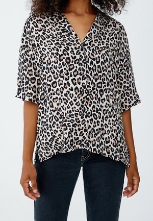 Femme portant un chemisier à manches courtes imprimé léopard avec décolleté en V et jeans foncés, debout devant un fond blanc.