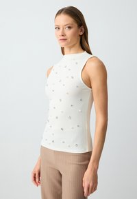 Top bianco a costine senza maniche con scollatura alta, decorato con vari dettagli in argento e perle, abbinato a pantaloni beige a righe.