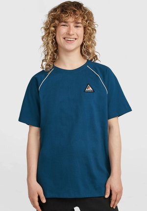 Persoon met krullend blond haar, gekleed in een blauw T-shirt met korte mouwen met een klein driehoekig logo op de borst, glimlachend en staand tegen een effen achtergrond.