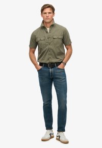 Camisa de botones de manga corta en color verde oliva con bolsillos en el pecho, combinada con jeans azules y zapatillas blancas con detalles en negro. Fondo plano.