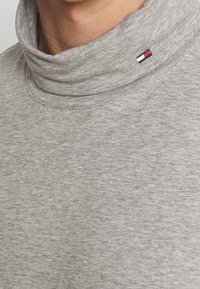 Grau, weicher Baumwollrollkragenpullover mit hohem Kragen, sichtbaren Nähten und einem kleinen Logo-Label in Rot, Weiß und Blau am Kragen.