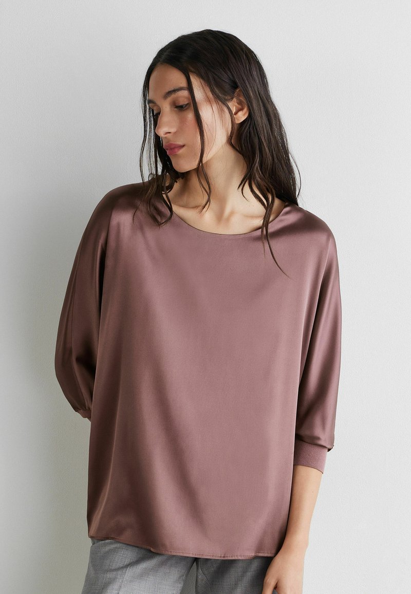 Frau mit langen dunklen Haaren, die eine lockere mauve Satinbluse mit dreiviertel Ärmeln trägt und vor einer schlichten weißen Wand steht.