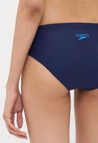 Navy blå svømmebukser laget av glatt, elastisk materiale; har et Speedo-logo i lyseblått, med lav midje-design og minimale sømdetaljer.