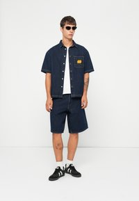 Camisa de mezclilla oscura y pantalones cortos, ambos con un ajuste relajado. La camisa cuenta con un bolsillo en el pecho con una etiqueta amarilla. Gafas de sol negras y zapatillas deportivas.