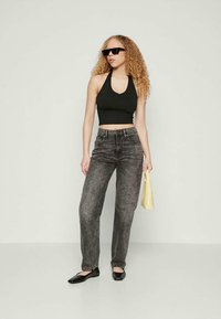 Donna con capelli ricci che indossa un top corto nero a collo alto, jeans neri sbiaditi, ballerine nere, occhiali da sole, mentre tiene una piccola borsa gialla contro un muro bianco.