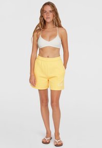Shorts di cotone gialli con vita elastica, con testo bianco. Abbinati a un top bikini bianco e infradito su uno sfondo neutro.