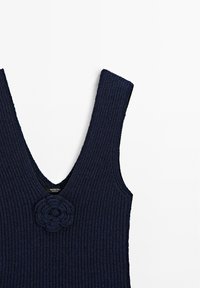 Námořnický pletený top bez rukávů s hlubokým výstřihem do V, širokými rameny a květinovým prvkem na přední straně. Texturovaná látka s žebrovaným designem.