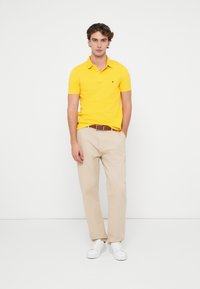 Gul bomull polo skjorte, beige chinos og hvite sneakers. Poloshirten har krage og en liten logo på brystet. Glatt tekstur.