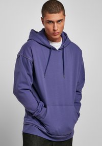 Lágy anyagból készült lila kapucnis jumper kenguruzsebbel, állítható húzózsinórral és bordázott mandzsettával. Lazább fazonnal és minimális varrási részletekkel rendelkezik.