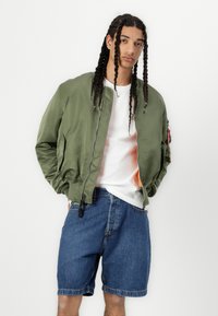 Veste bomber vert olive avec fermeture à zip, poches latérales et doublure colorée, portée avec un short en jean bleu. T-shirt blanc en dessous.