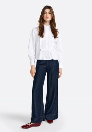 Camicia bianca a blouson con pieghe verticali e colletto alto, abbinata a pantaloni in denim scuro a gamba larga e ballerine bordeaux.