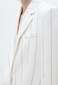 Blazer blanc à fines rayures verticales noires, col à revers cranté et poche poitrine sur une personne à la peau claire visible du cou jusqu'au milieu du torse.