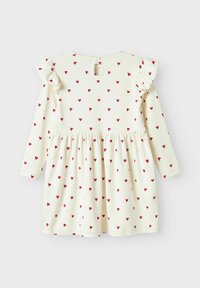 Robe à manches longues en crème avec un motif de cœurs rouges. Épaules volantées et fermeture en forme de clef au dos. Fabriquée en tissu doux et léger.