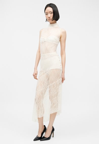 Γυναίκα που φορεί ένα άσπρο διάφανο δαντελένιο φόρεμα χωρίς μανίκια με ενσωματωμένα σορτς και μαύρα παπούτσια με ψηλό τακούνι, στεκόμενη μπροστά σε απλό φόντο.