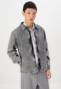 Gray denim jacket with a collared neck, snap buttons, and elastic cuffs. Worn over a white checkered shirt. Clean design with a subtle fade.