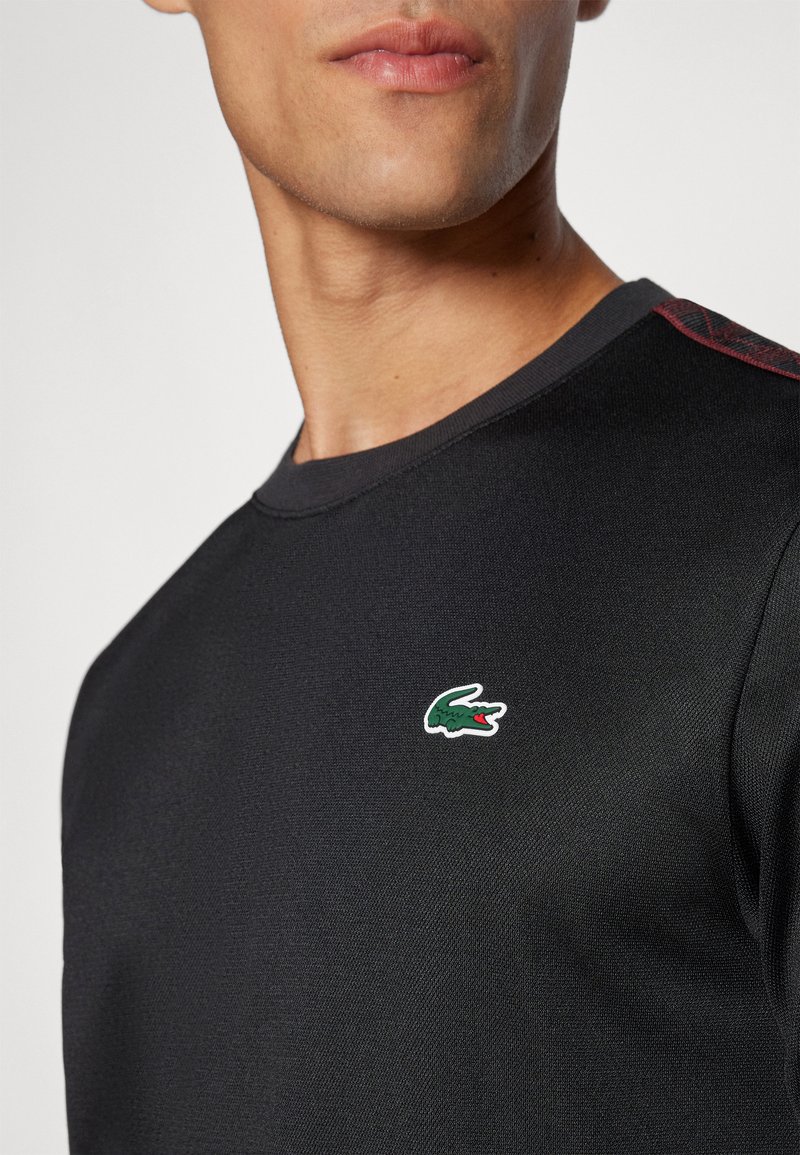 Sweatshirt noir à col rond en tissu texturé, présentant un petit logo Lacoste vert et rouge sur la poitrine gauche. Accents rouges sur les coutures des épaules.