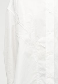Λευκό πουκάμισο με κουμπιά, διακοσμημένο με λεπτό κεντητό λουλουδάτο σχέδιο στην αριστερή πλευρά, εστιασμένο σε λεπτομερή κέντημα και υφή υφάσματος.