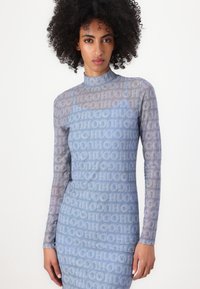 Langere jurk met lange mouwen in lichtblauw, gemaakt van transparant materiaal, met een herhalend logopatroon in donkerder blauw. Hoge halsontwerp, aansluitende silhoutte.