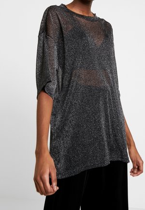 Persona vestida con una blusa negra suelta y translúcida con detalles de purpurina plateada y mangas cortas, combinada con pantalones negros, sobre un fondo liso.