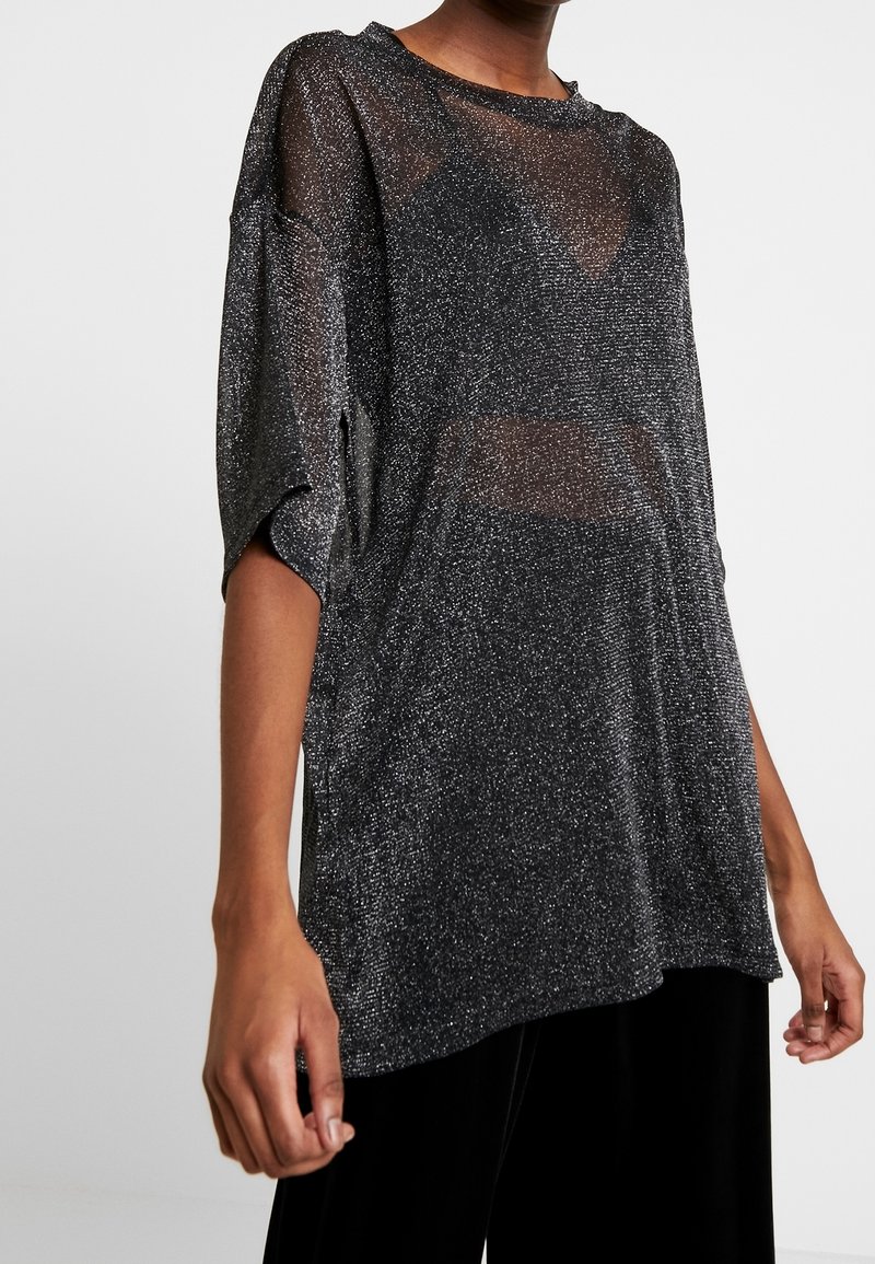 Personne portant un haut noir ample et transparent avec des détails scintillants argentés et des manches courtes, associé à un pantalon noir, sur un fond uni.