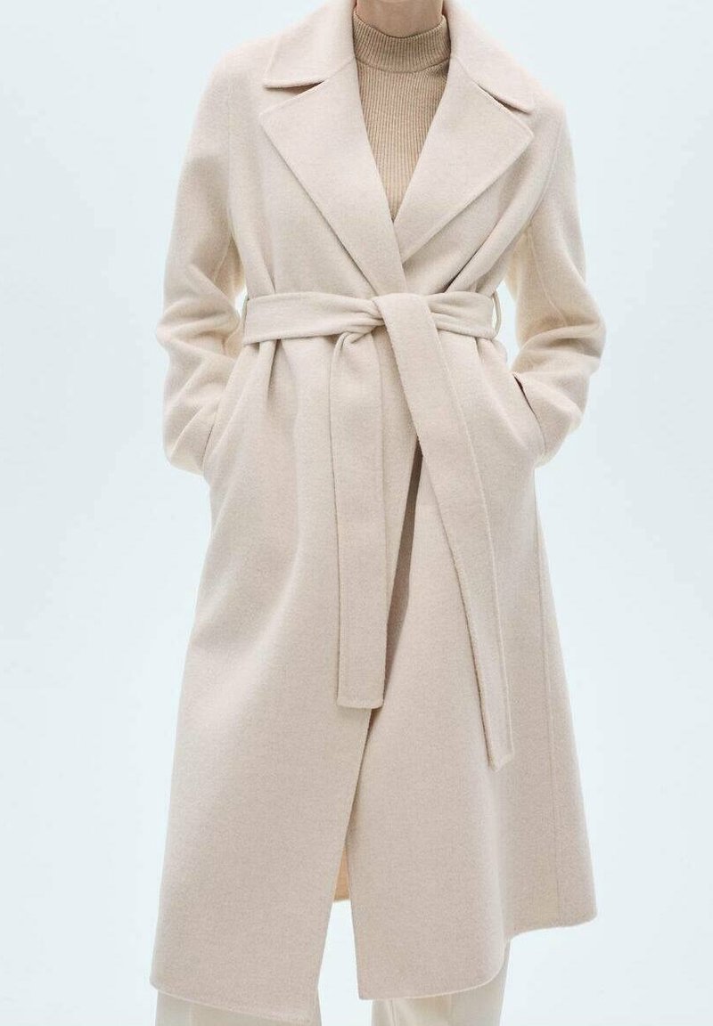 Femme portant un manteau en laine beige clair ceinturé par-dessus un pull à col roulé marron, mains dans les poches du manteau, debout devant un fond uni.
