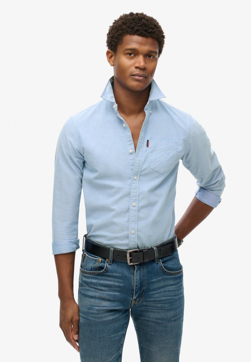 Lichtblauw overhemd met lange mouwen en knoopsluiting, zak op de linkerborst, gedragen met donkerdenim jeans en een zwarte riem.